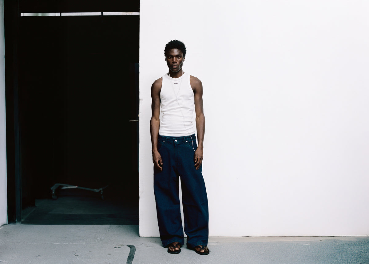 REAMO – model in Sayeem raw denim and a white tank top standing against a white wall next to a dark doorway